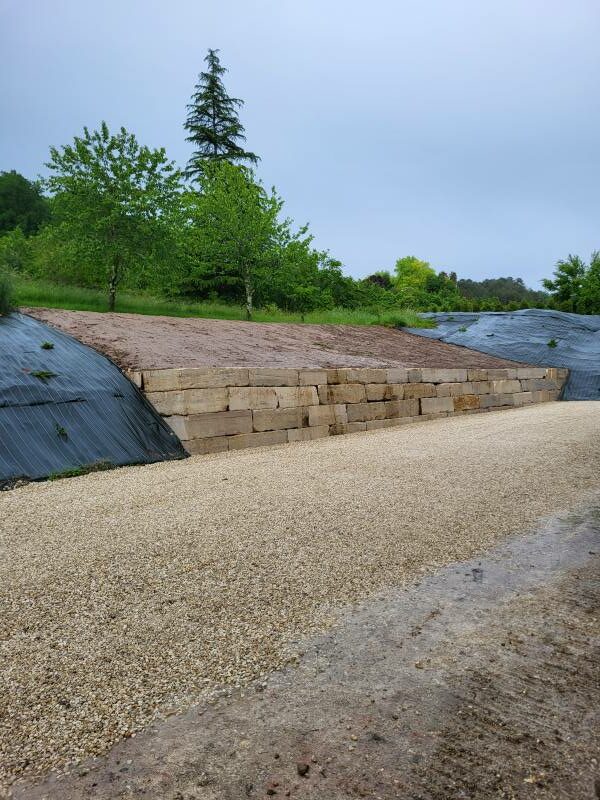 Réalisation par l'entreprise SARL TCTP en Dordogne d'un enrochement en blocs de pierre naturelle de limeyrat pour l'aménagement d'un parking avec une finition en calcaire 16/22 après un éboulement de talus et remise en place de bâche de paillage sur les extrémités du mur.